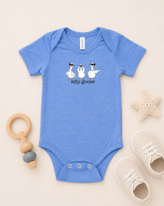 A baby short-sleeve bodysuit in heathered blue, laid flat on a light beige background. The bodysuit features a graphic of three white geese wearing sunglasses with the text 'silly goose' underneath.