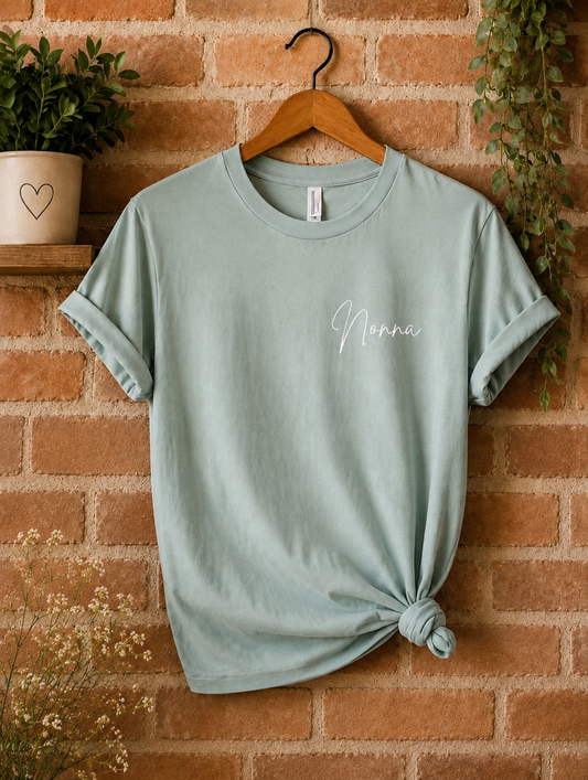 A sage green short-sleeve t-shirt with 'Nonna' printed in white script on the chest, hanging on a wooden hanger against a brick wall background with decorative plants.