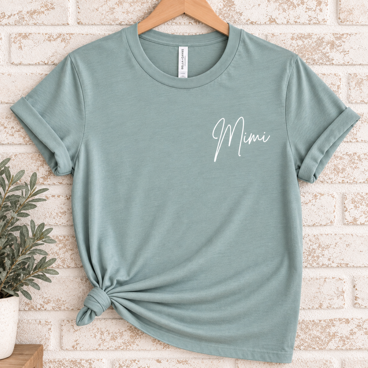 A sage green short-sleeve t-shirt with 'Mimi' written in white script on the chest, styled on a wooden hanger with a knot at the bottom hem against a light brick wall background.