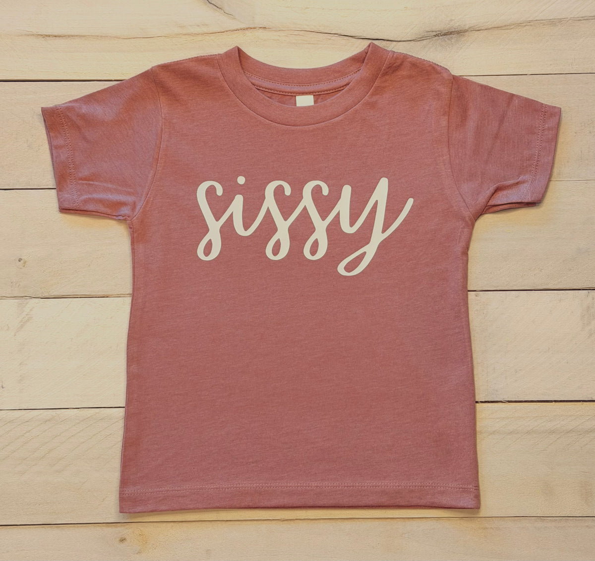 Children's heather mauve t-shirt with 'sissy' in cursive printed on it, placed on a wooden surface.