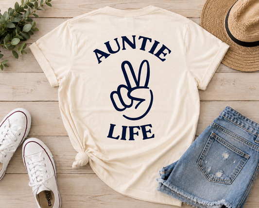 A cream-colored graphic t-shirt featuring the text 'AUNTIE LIFE' in navy blue and a peace sign hand graphic, styled in a flat lay with white sneakers, denim shorts, a straw hat, and green leaves on a light wood background.