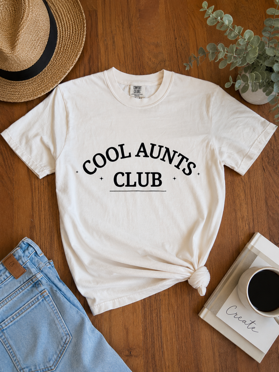 An ivory graphic t-shirt with 'COOL AUNTS CLUB' printed in black arched text, styled in a flat-lay on a wooden table with a straw hat, jeans, a plant, and a coffee mug.