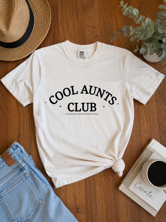 An ivory graphic t-shirt with 'COOL AUNTS CLUB' printed in black arched text, styled in a flat-lay on a wooden table with a straw hat, jeans, a plant, and a coffee mug.