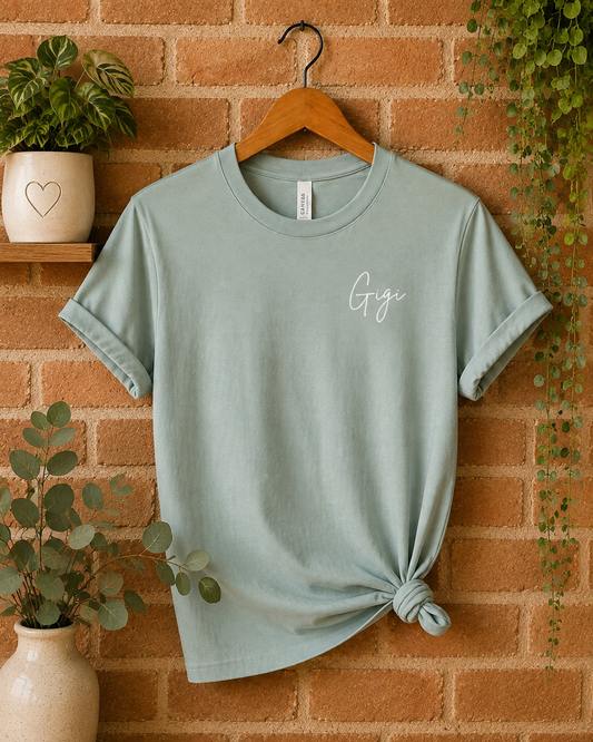 A sage green short-sleeve t-shirt with a small white script 'Gigi' logo on the chest, styled on a wooden hanger against a brick wall with plants.