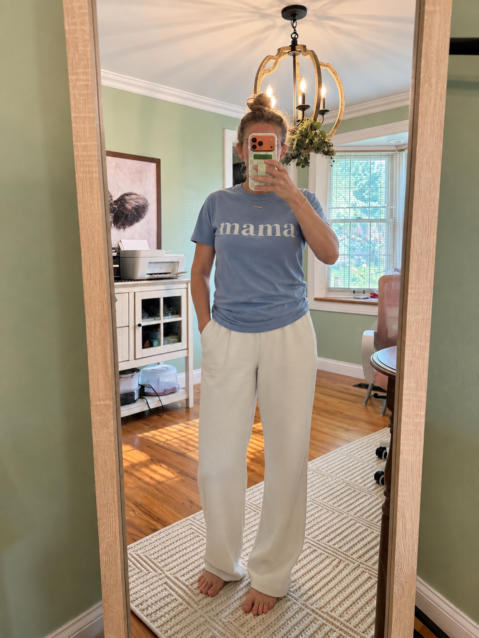 A woman takes a mirror selfie in a home setting, wearing a light blue t-shirt with the word 'mama' printed in white across the chest, paired with off-white wide-leg pants.