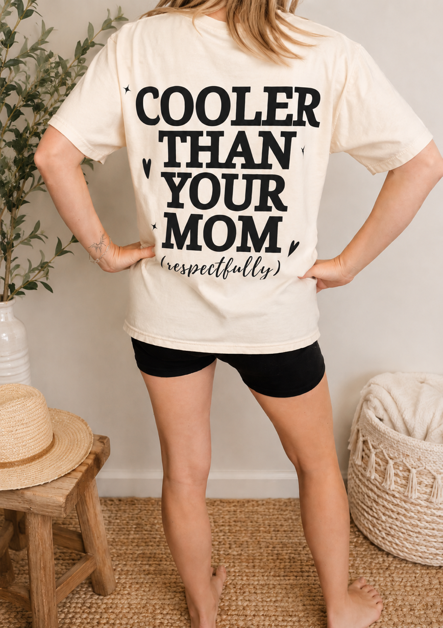 A woman stands with her back to the camera wearing an ivory-colored graphic t-shirt that reads 'COOLER THAN YOUR MOM (respectfully)' in bold black text, paired with black athletic shorts. The setting is a cozy indoor room with a wooden stool, a straw hat, a plant, and a woven basket.