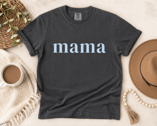 A dark charcoal gray t-shirt with the word 'mama' printed in a white serif font across the chest, styled in a cozy flat-lay arrangement with a coffee mug, woven hat, and neutral textiles.