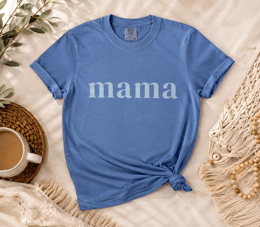 A blue short-sleeve t-shirt with the word 'mama' printed in white across the chest, styled in a flat lay on a beige fabric background with a cup, woven mat, and macrame decor.