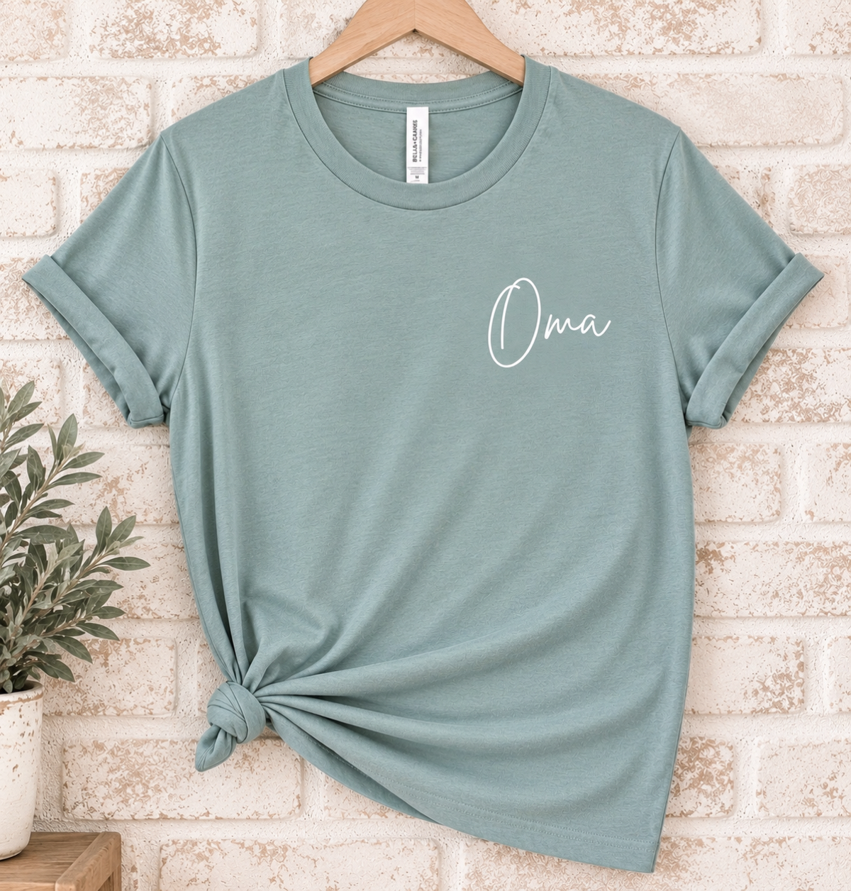 A sage green short-sleeve t-shirt with a white script 'Oma' graphic on the chest, styled on a wooden hanger against a light brick wall with a plant in the corner.
