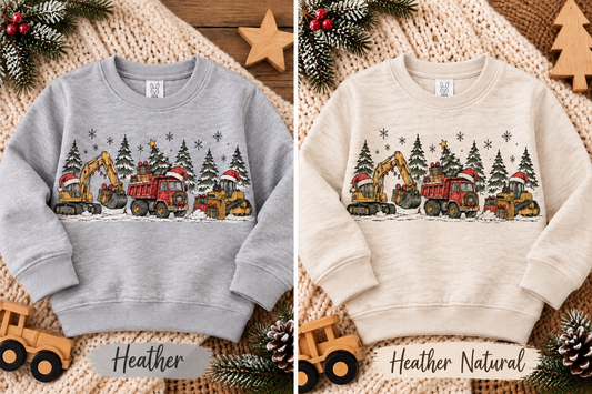 Two children's crewneck sweatshirts shown side by side on a cozy knit blanket with holiday pine branches and wooden toys. The left sweatshirt is heather gray and labeled 'Heather'; the right is heather natural cream and labeled 'Heather Natural'.