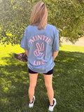 Person wearing a blue jean t-shirt with pastel pink 'Auntie Life' printed on the back, standing outdoors.