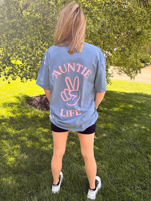 Person wearing a blue jean t-shirt with pastel pink 'Auntie Life' printed on the back, standing outdoors.