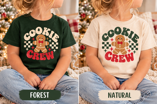 Two children seated cross-legged wearing matching holiday graphic T-shirts, shown side by side to display two color options. The left child wears a forest green shirt with a gingerbread cookie graphic and the words 'COOKIE CREW' in cream, red, and green.