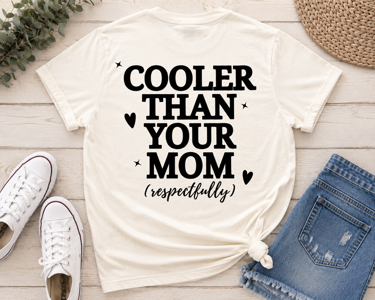 An ivory graphic t-shirt with the text 'COOLER THAN YOUR MOM (respectfully)' in black, styled in a flat lay with blue jeans, white sneakers, a straw hat, and greenery on a light wood background.