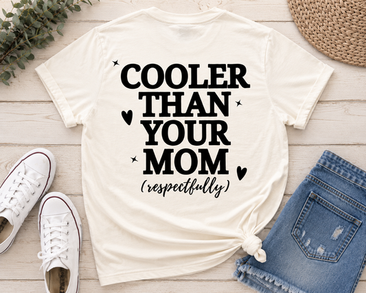 An ivory graphic t-shirt with the text 'COOLER THAN YOUR MOM (respectfully)' in black, styled in a flat lay with blue jeans, white sneakers, a straw hat, and greenery on a light wood background.
