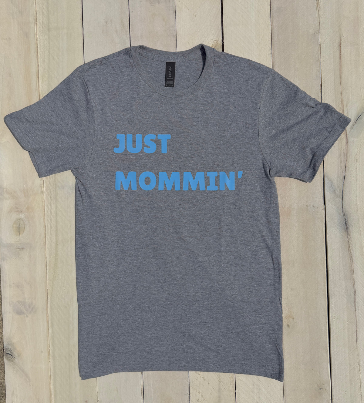 Gray t-shirt with blue text 'JUST MOMMIN'' on a wooden surface