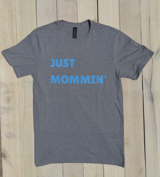 Gray t-shirt with blue text 'JUST MOMMIN'' on a wooden surface