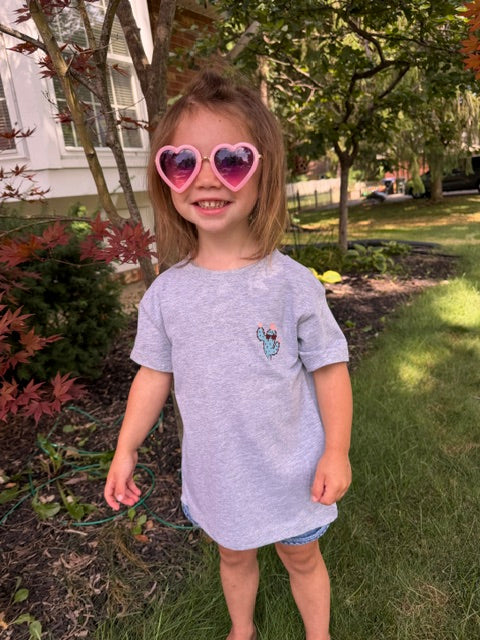 A young girl wearing a Heather gray  t-shirt with a cactus wearing shades graphic on the front left chest. She has on denim shorts while standing outdoors on a grassy lawn, smiling and wearing heart-shaped pink sunglasses.