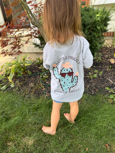 A child wearing a heather gray t-shirt with a cartoon cactus graphic and the text 'A Little Prickly, A Lot of Sweet' while standing outdoors on a grassy lawn.