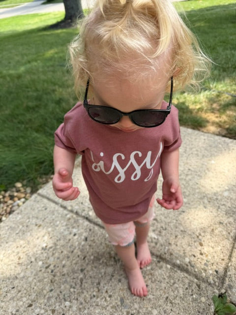 A toddler standing outdoors on a concrete path wearing a heather mauve short-sleeve t-shirt with 'sissy' printed in white script, light pink shorts, and black sunglasses.